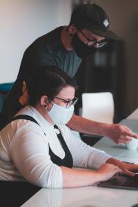 A man leans over a woman who is working on a laptop. They are both wearing face masks