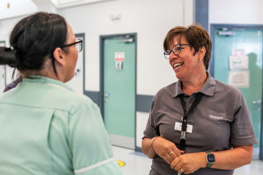 Two women wearing glasses are talking in a hallway and smiling