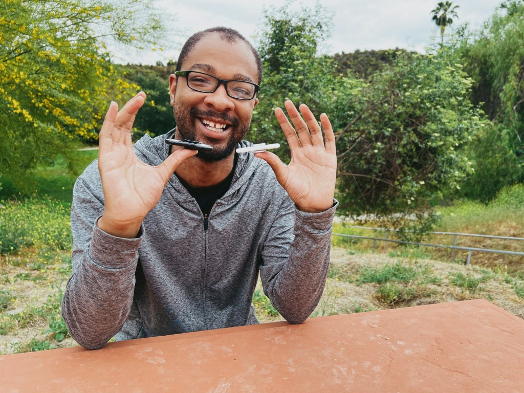 A man sits at a table outdoors. He wears glasses and is smiling, as he balances a pen on both thumbs