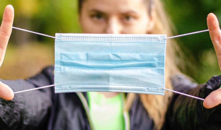 Close up of a surgical face mask. In the background is a white woman who is stretching the mask taut
