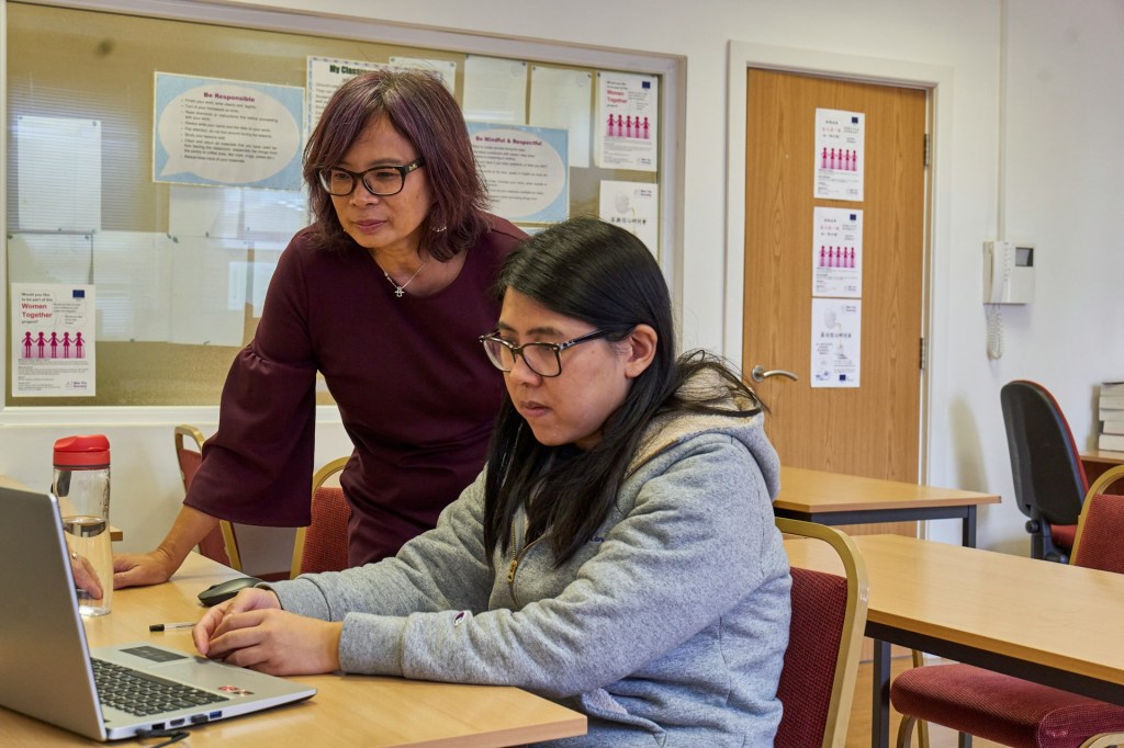 A woman stands over another woman who is working on a computer