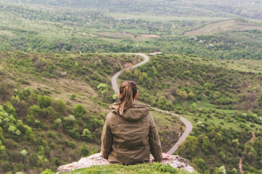 Woman sitting on a cliff edge, looking over a vast countryside, with a road winding into the distance