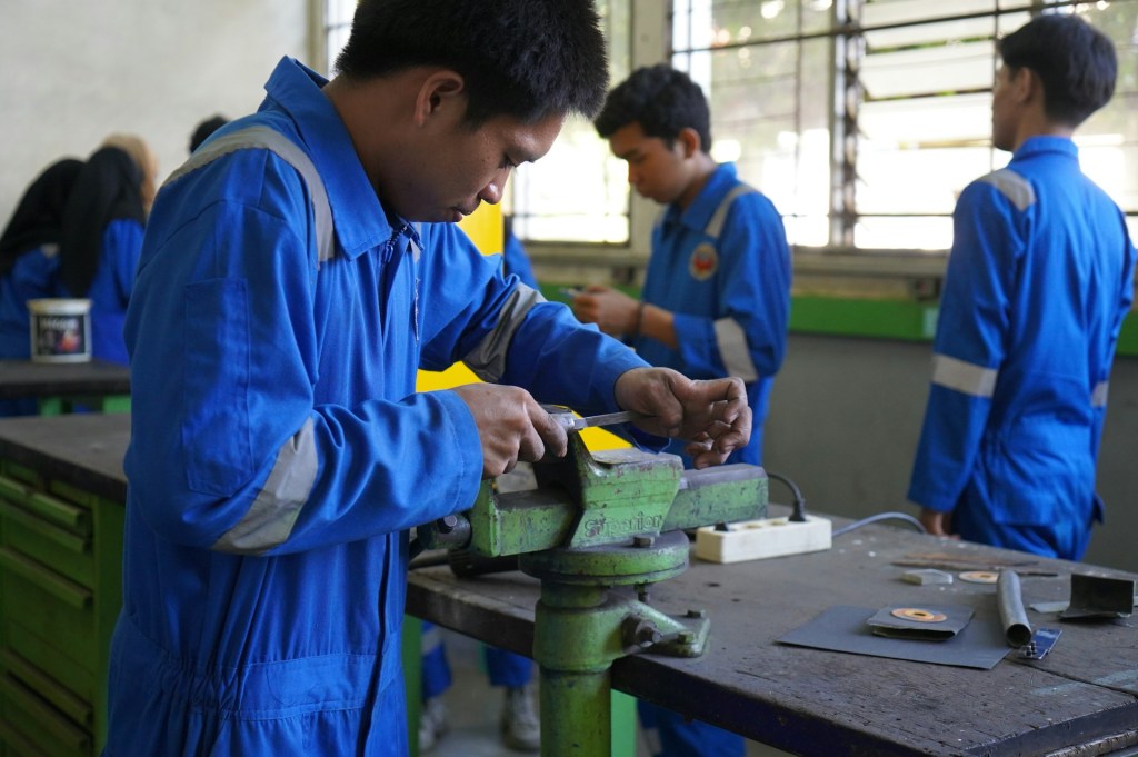 A young man in a blue uniform is working with metal in a clamp, while other people in the same uniform stand in groups in the background
