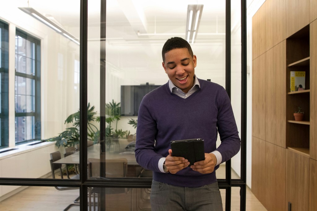 Inside an office, a man smiles as he reads a tablet