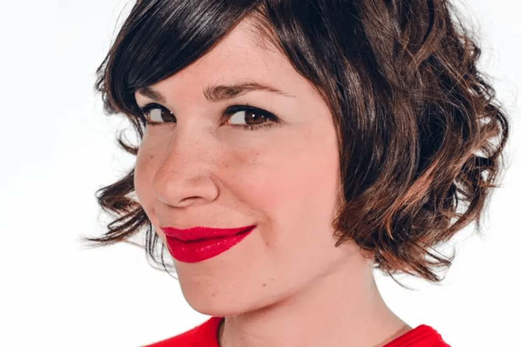 Carrie Brownstein is a white woman with short brown, curly hair. She wears red lipstick and smiles