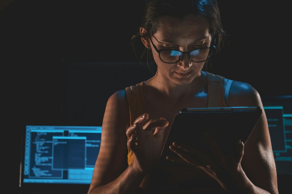 A woman works on a tablet in a dark room, with a computer in the background