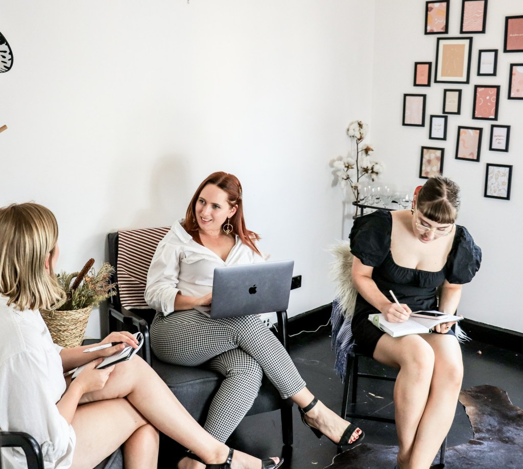 Three women sit in an office, two are talking, one is on her laptop, and another takes notes