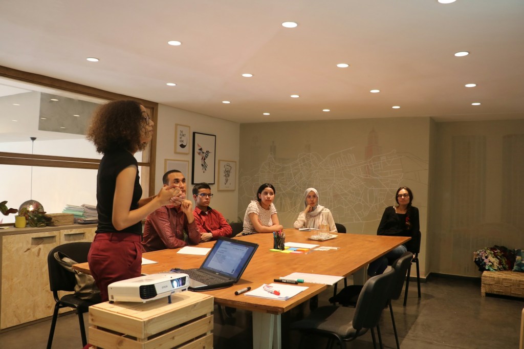 A committee sitting around a large table watches a woman give a presentation
