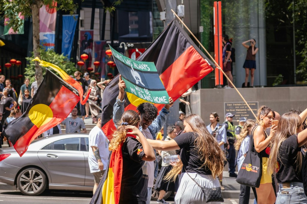 Young people at a march hold Aboriginal flags, while another young person holds a sign painted like the Torres Strait Islander flag, with the words: 'Always was, will be my Island home'