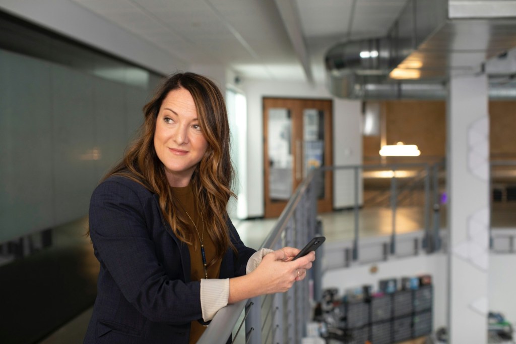 A woman holds a phone over a balcony, she smiles as she looks to the side
