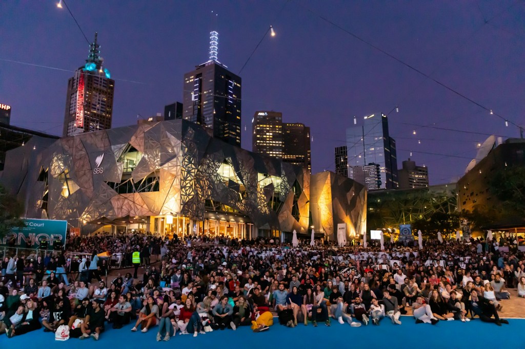 A large crowd sits on the ground at Federation Square, Melbourne, at night