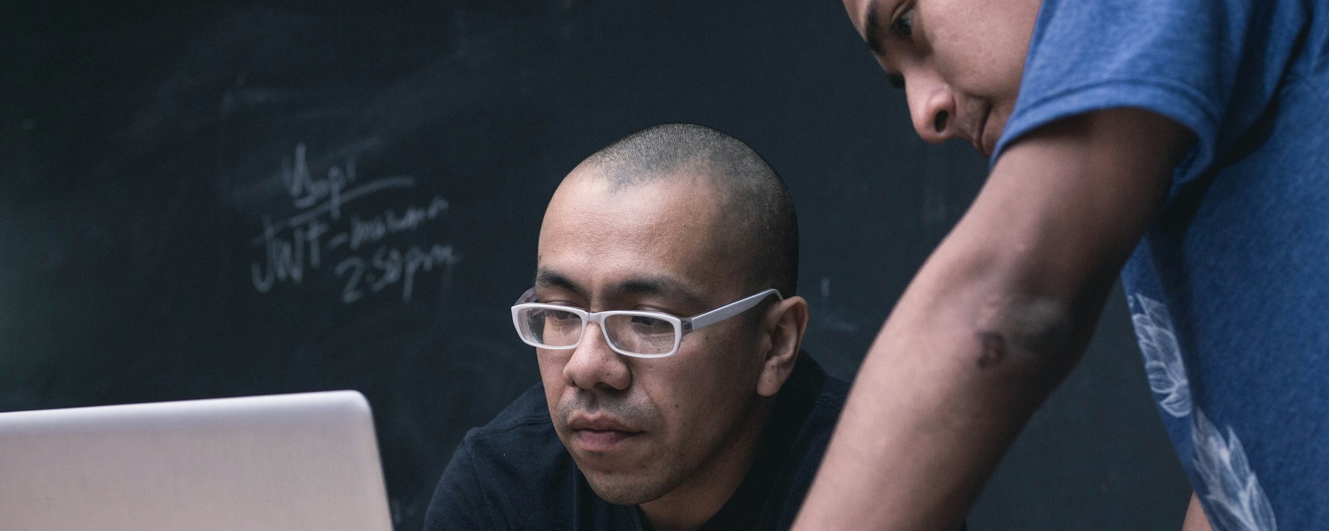 A Black man with white glasses sits in front of his laptop, while a Latin man stands over him looking at the screen