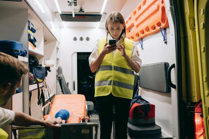 A white woman paramedic stands inside an ambulence, reading her phone