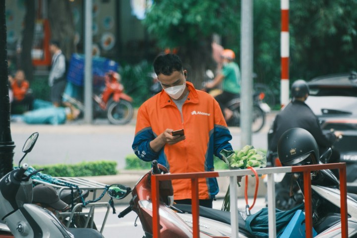 Asian man wearing a face mask reads his phone on the street beside his motorbike
