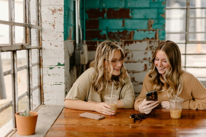Two young white women are smiling while looking at one of their phones, in a cafe
