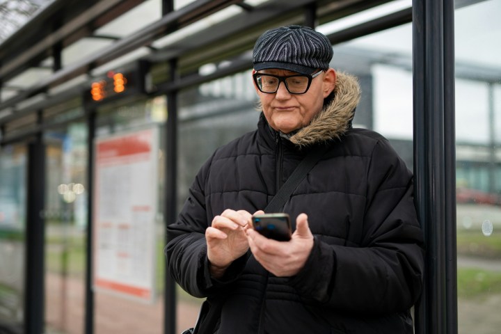A white older man in a hat and jacket writes on his phone outdoors