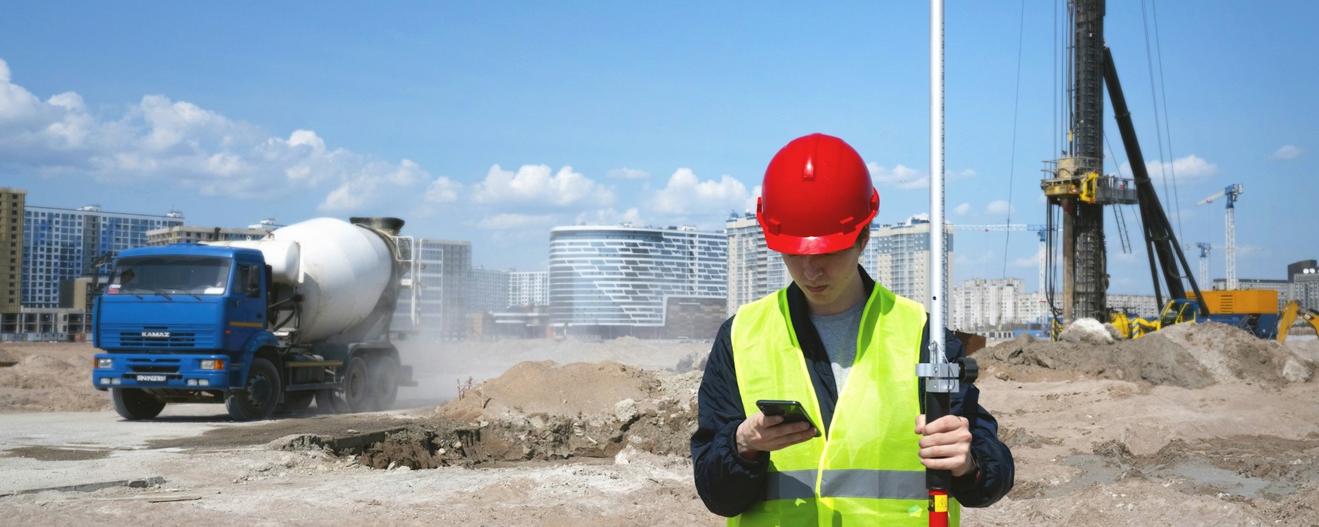 Young male construction worker looks at his phone while holding a long pole. He wears a hardhat and hi-vis vest. A cement truck and mounds of dirt are in the background