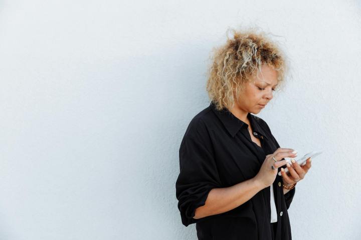 Black woman with curly blonde hair reads her mobile phone outdoors against a white wall