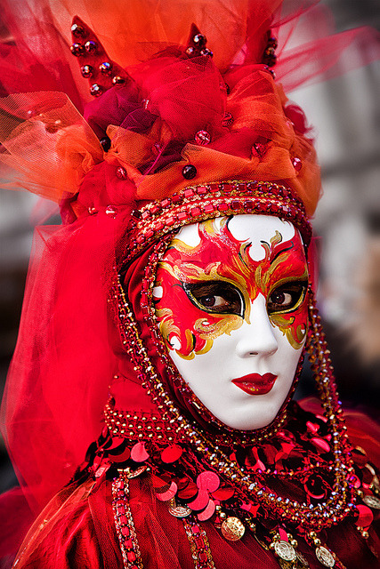 Person wearing a white mask with red and gold swirls, and a red costume with a high headdress with beads from the head through the collar