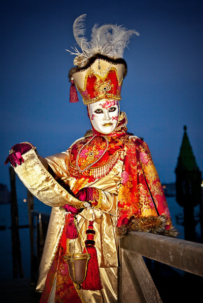 Person wearing a white mask with a tall mitre hat, and gold and red ceremonial costume
