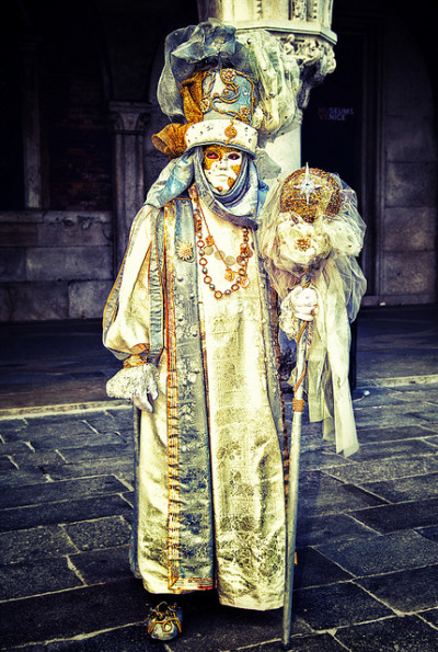 Person wearing a green and gold costume with light blue flourishes, including a white and orange mask and a sceptre