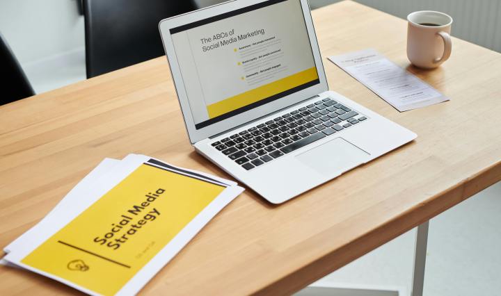 A laptop on a table, open to a webapge that reads, 'The ABCs of social media marketing.' A cup of coffee is on the right, and on the left is a print out saying, 'Social media strategy'