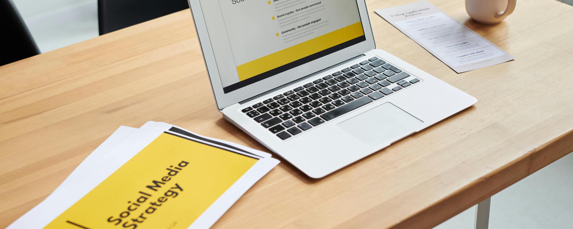 A laptop on a table, open to a webapge that reads, 'The ABCs of social media marketing.' A cup of coffee is on the right, and on the left is a print out saying, 'Social media strategy'