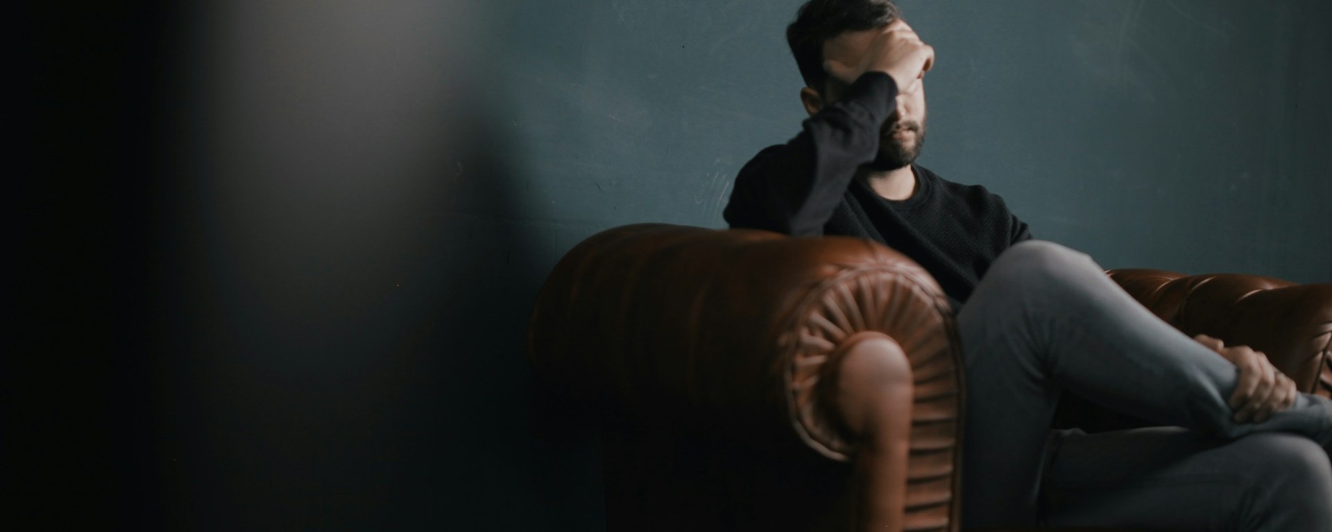 White man sits on a leather couch with his hand covering his eyes