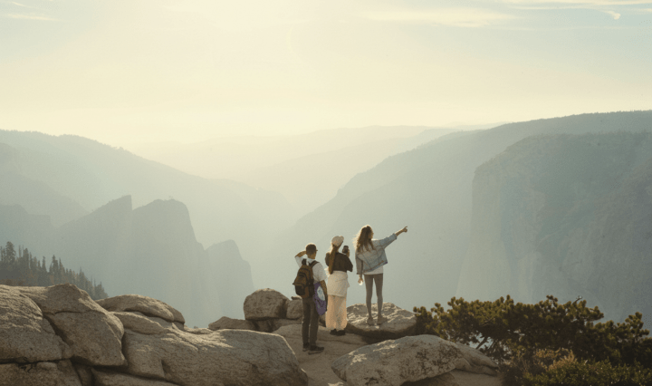Two white women and a man stand at the edge of a cliff. One of them points into the distancce while the other takes a photo with her phone