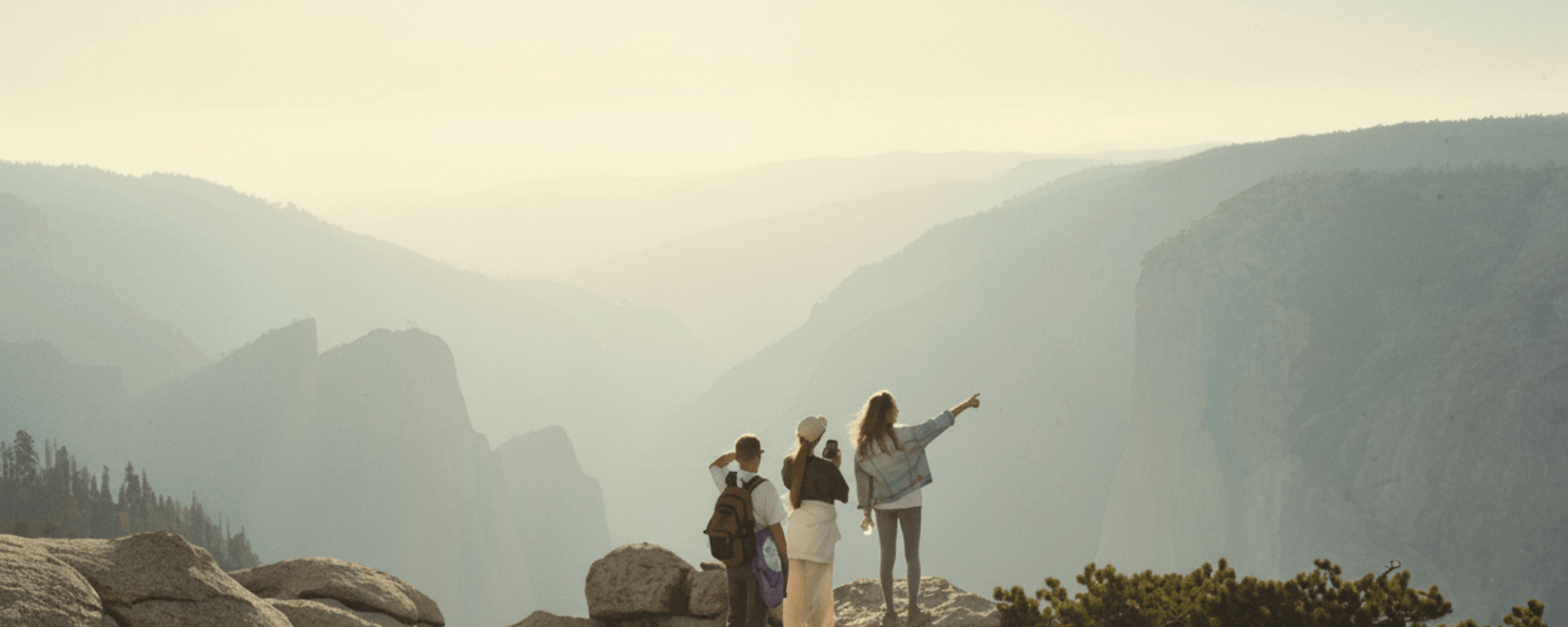 Two white women and a man stand at the edge of a cliff. One of them points into the distancce while the other takes a photo with her phone