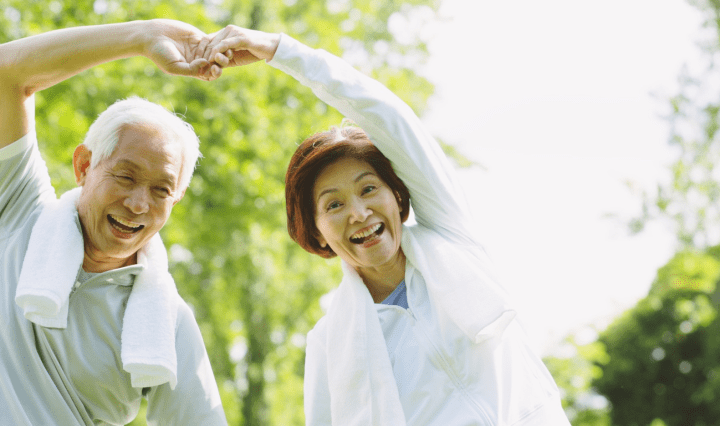 An older Asian man and woman exercise in the park. They arch their arms and hold hands in the air