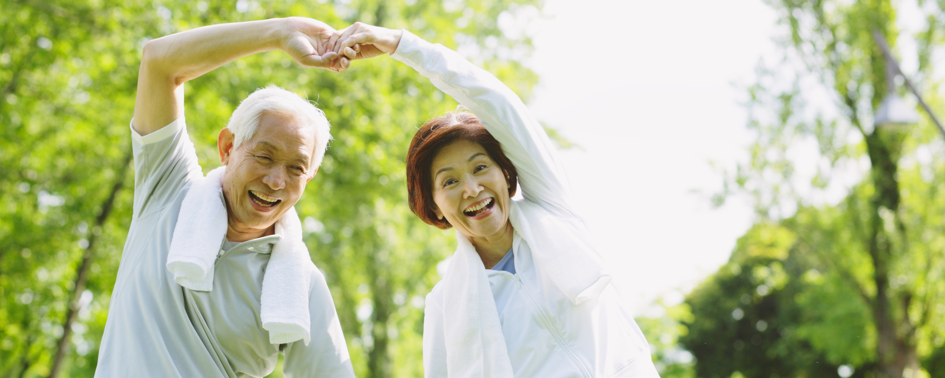 An older Asian man and woman exercise in the park. They arch their arms and hold hands in the air