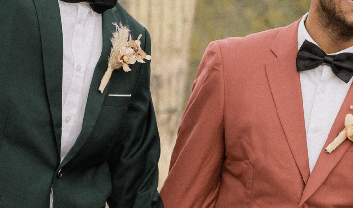 Two grooms stand side by side holding hands. One wears a pink suit and the other a black suit