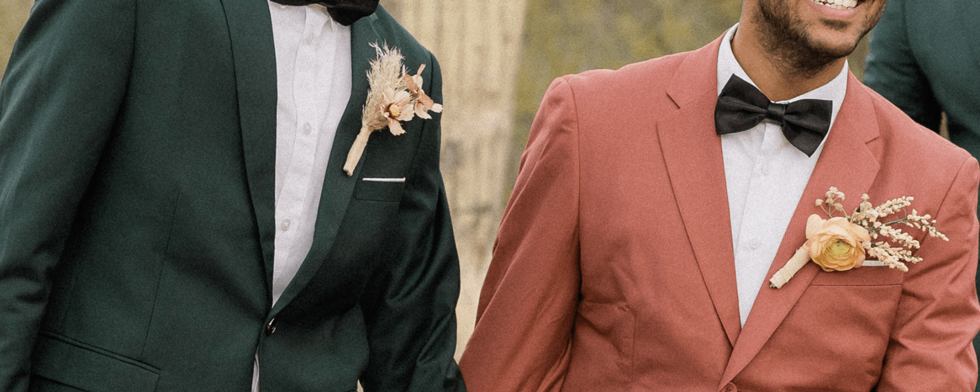 Two grooms stand side by side holding hands. One wears a pink suit and the other a black suit