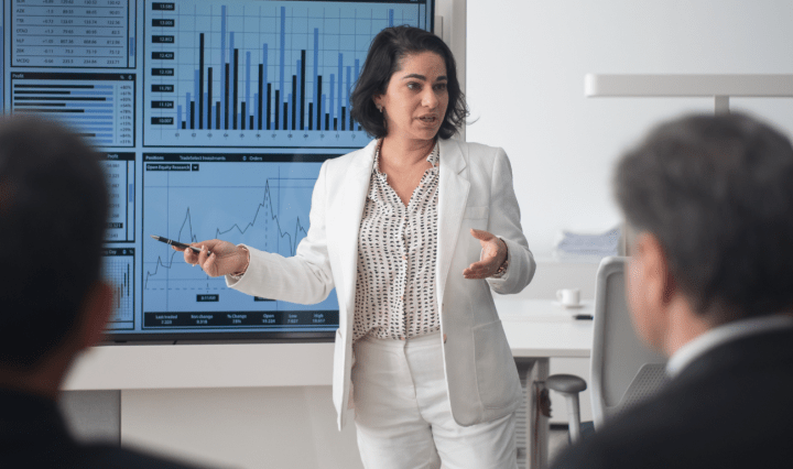 A Middle Eastern woman presents charts to a crowd of men in dark suits