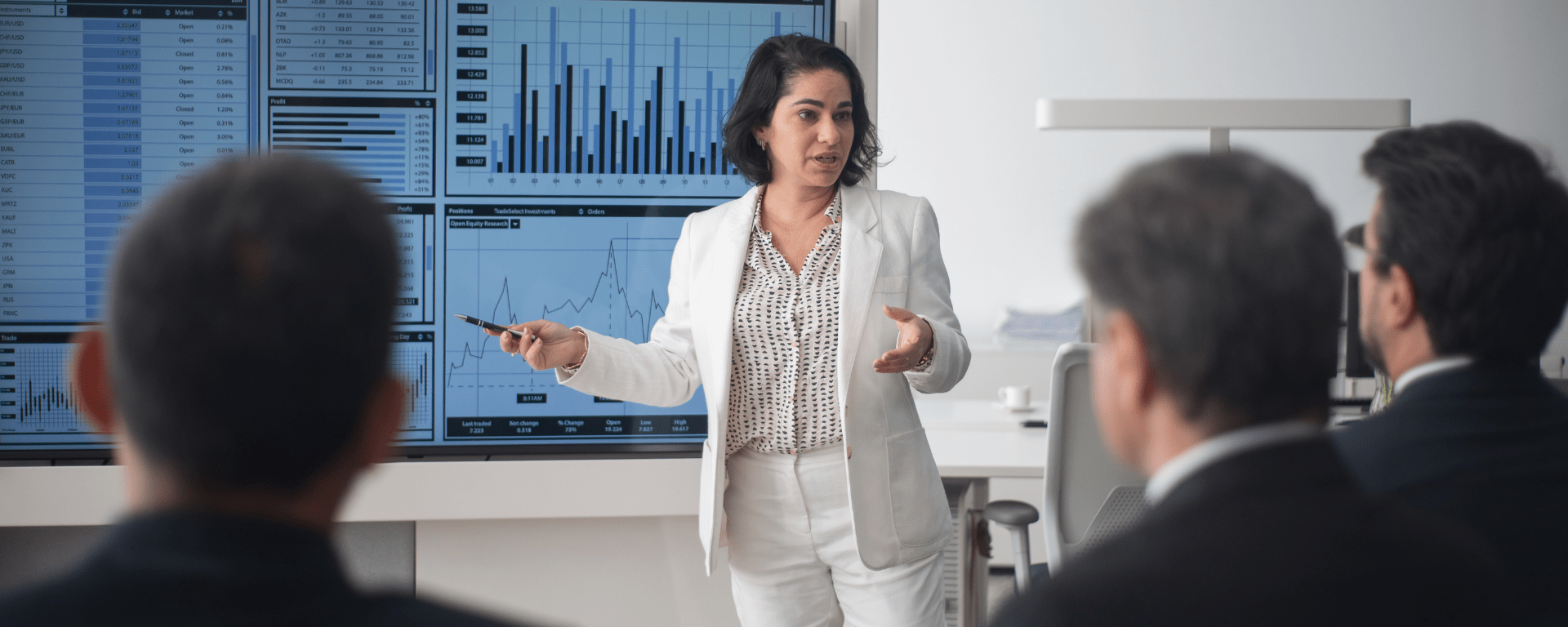 A Middle Eastern woman presents charts to a crowd of men in dark suits