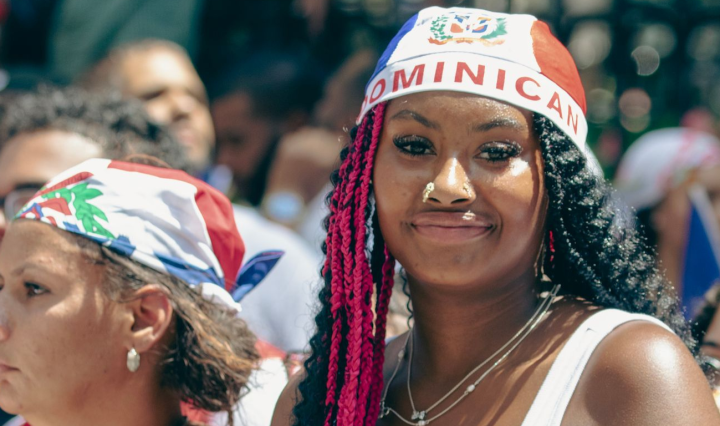 Black Dominican women are in a crowd. The woman in the foreground wears a flag as a cap, with the word "Dominican" on the front
