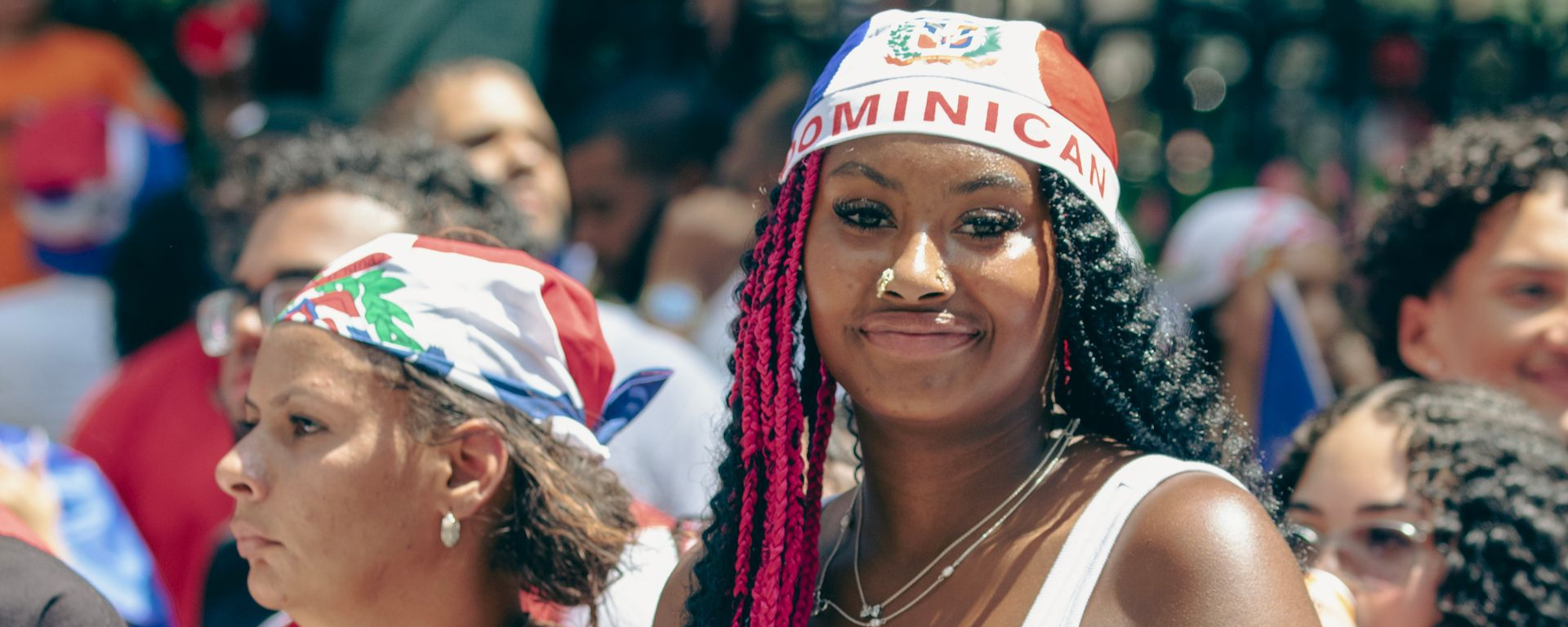Black Dominican women are in a crowd. The woman in the foreground wears a flag as a cap, with the word "Dominican" on the front