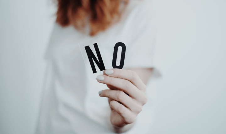 A white woman blurred in the background holds up two letters close to the camera that say: N O
