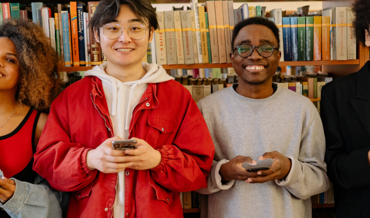Four racially diverse students stand in front of book shelves in a library. They smile and hold their phones