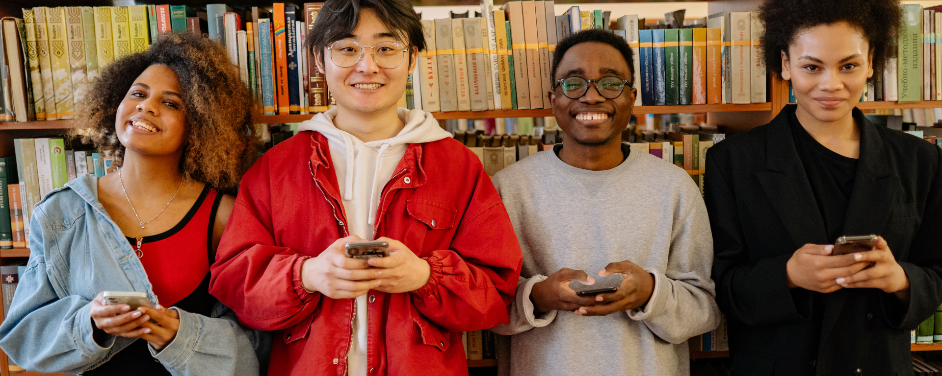 Four racially diverse students stand in front of book shelves in a library. They smile and hold their phones
