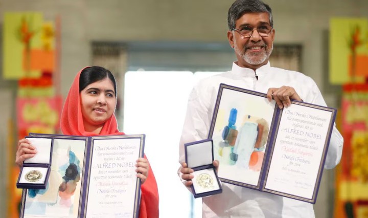 Malala Yousafza and Kailash Satyarthi stand side by side, holding up their medals and certificate. Yousafza is a Pakistani girl with black hair parted to the side, covered in a red hijab. Satyarthi is an Indian man with grey hair and glasses