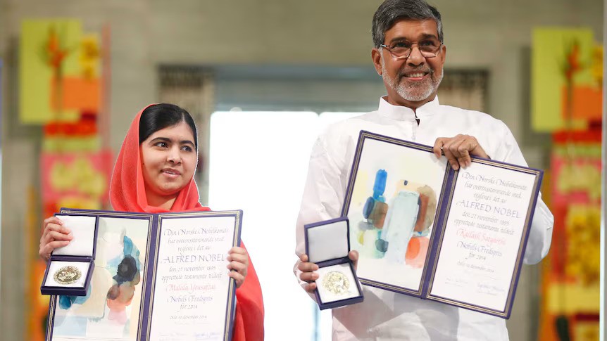 Malala Yousafza and Kailash Satyarthi stand side by side, holding up their medals and certificate. Yousafza is a Pakistani girl with black hair parted to the side, covered in a red hijab. Satyarthi is an Indian man with grey hair and glasses