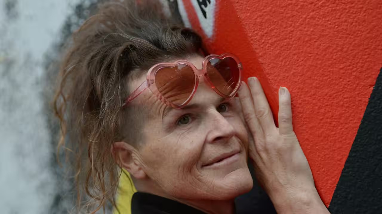 Norrie is a trans woman who has wavy brown hair. She wears pink heart-shaped sunglasses and hugs a wall, as she smiles