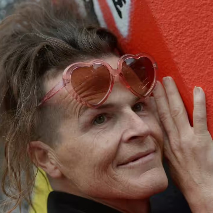 Norrie is a trans woman who has wavy brown hair. She wears pink heart-shaped sunglasses and hugs a wall, as she smiles