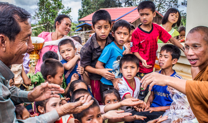 A man and children hold their hands out to a Buddhist monk in an outdoor area