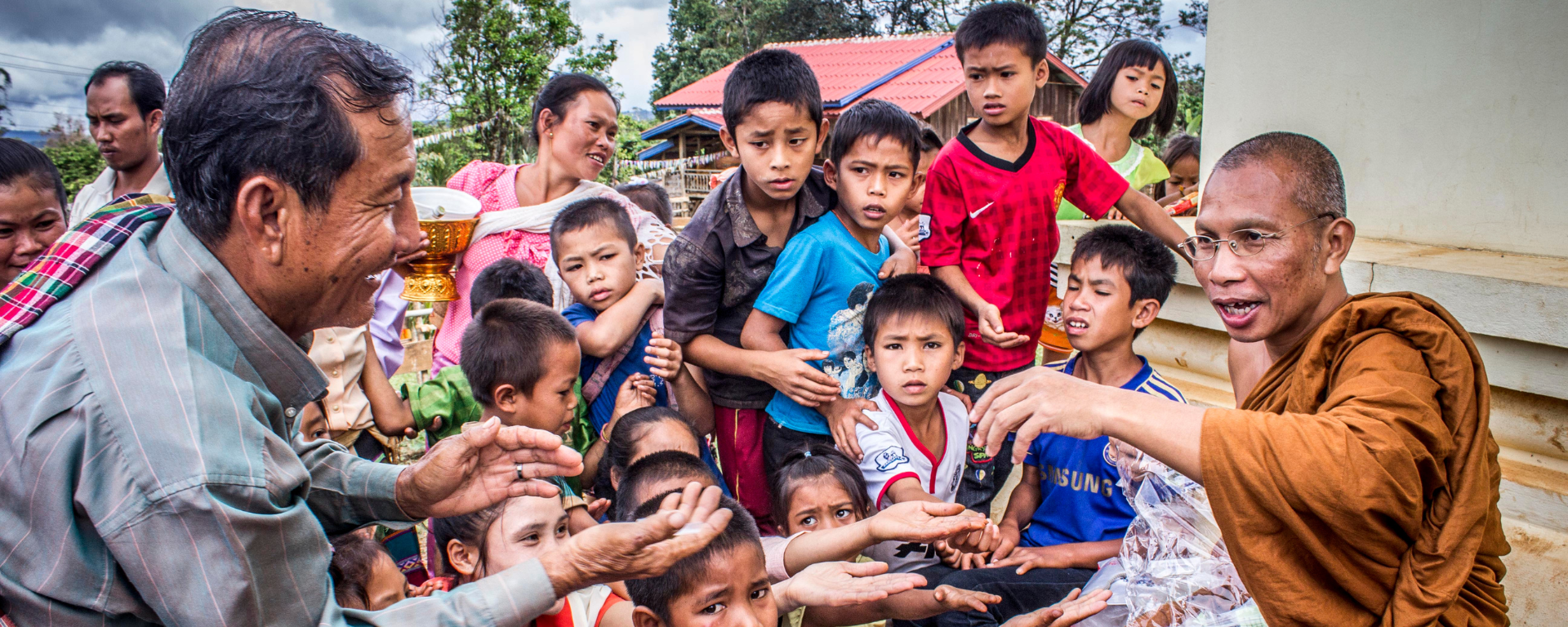 A man and children hold their hands out to a Buddhist monk in an outdoor area
