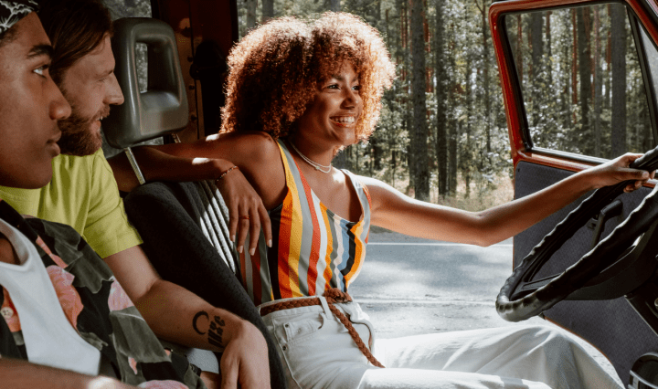A smiling young Black woman sits with the driver's door open, while a Black man and white man look at her