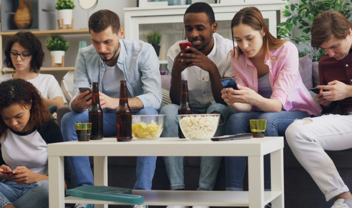 A diverse group of friends sit on a couch with snacks and drinks on a table, as they each look at their phones