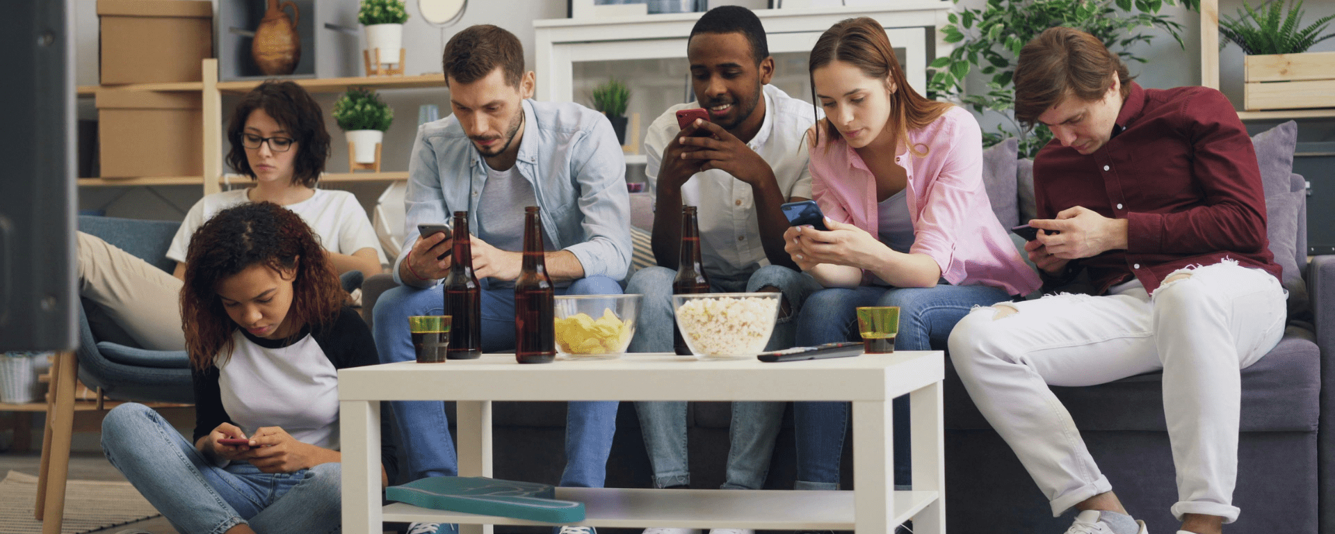 A diverse group of friends sit on a couch with snacks and drinks on a table, as they each look at their phones